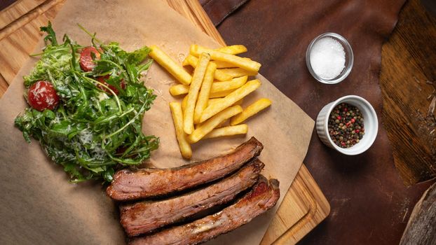 Beef rib with French fries and arugula salad with cherry tomatoes and parmesan cheese served on board covered with food grade paper. Restaurant concept.