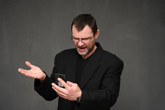 Caucasian man in a black jacket with glasses looks displeased at the phone with emotions on a gray background