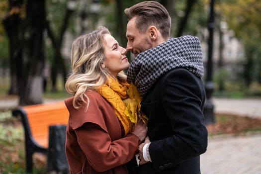 Passionate couple handsome guy hold his girlfriends hands and touching noses stand in fall town park wearing coats and scarfs. Smiling couple hugging in autumn park.