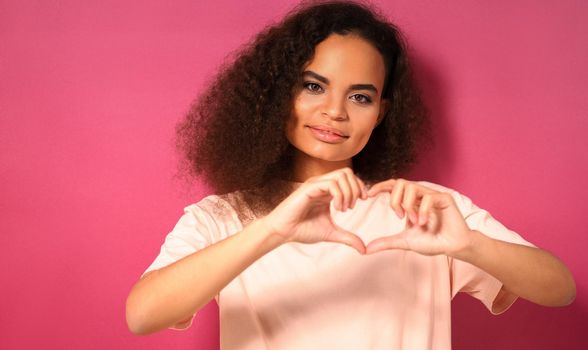 Love gesture with a tender look beautiful young African American woman looking positively at camera wearing peachy t-shirt isolated on pink background. Beauty concept.