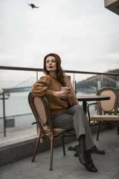 Lovely french young woman sitting at restaurant terrace with coffee mug looking at camera. Portrait of stylish young woman wearing autumn coat and red beret outdoors.