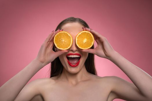 Funny beautiful beauty model young woman holding two juicy orange slices as her eyes or glasses. Charming joyful funny lady with red lips and long hair isolated on pink background.