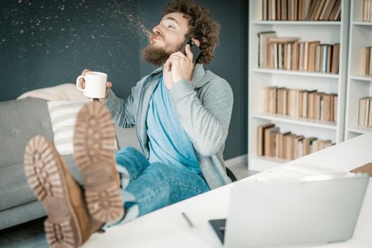 Surprised Man Hears the News on his Smartphone and Spits Out Coffee. Man has Legs on the Table and a Mug of Coffee in His Hand. Close-up. Bookshelves Background. High quality photo