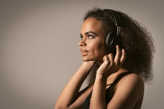 Capture away by music young African-American girl looking up away sideways listening music wearing headphones, isolated on grey background, emotionally move, have fun. Close up.