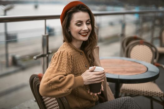 Happy young girl sitting outdoors street restaurant or terrace looking smiling away holding a coffee mug. Portrait of stylish young woman wearing autumn coat and red beret outdoors.