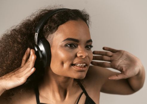 Young smiling african girl listening music in headphones with bare shoulders and long curly hair. Attractive hipster brunette girl enjoying free time isolated on grey background.