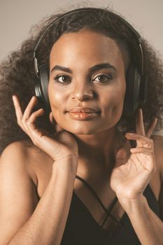 Young African-American girl listening music in headphones wearing black top isolated on grey background, emotionally move. Concept of emotions, facial expression, sales, ad, inclusion.