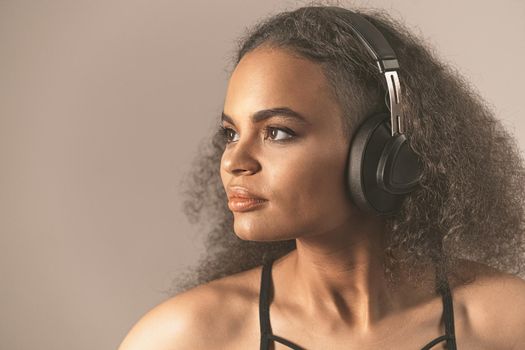 Beautiful young African-American girl listen music looking up away sideways wearing headphones, isolated on beige background. Charming girl with long curl hair listening music. Close up.