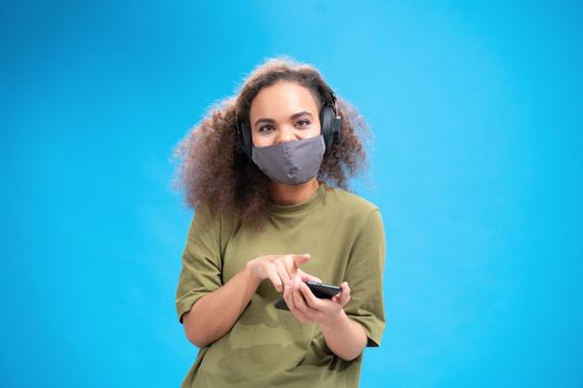 African american girl looking at camera wearing reusable face mask and listen music in headphones. Wearing safety mask while using smartphone to communicate on social distancing isolated on blue.
