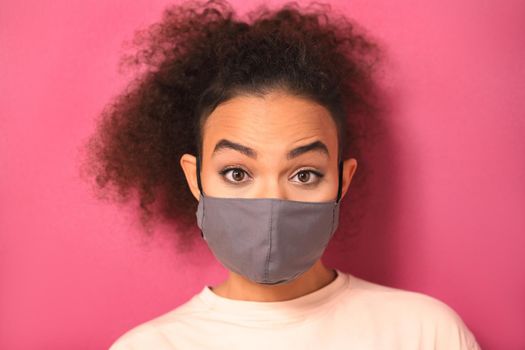 Recognize emotions in a reusable mask during quarantine African American girl in peachy t-shirt, to prevent others from corona COVID-19 and SARS cov 2 infection isolated on pink background.