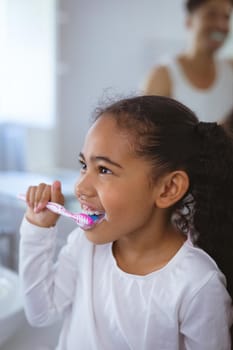 Multiracial girl brushing teeth while looking away with father in background. unaltered, family, togetherness and hygiene concept.