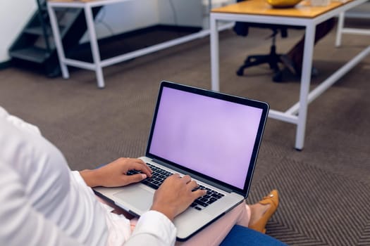 Low section of biracial young businesswoman using laptop with blank space while sitting in office. unaltered, creative business, copy space, modern office and wireless technology concept.