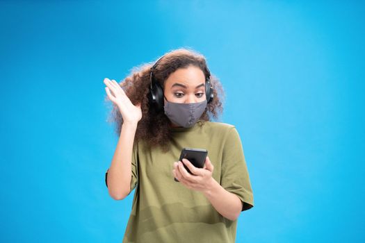 Gesturing african american girl looking at camera wearing reusable face mask and headphones. Wearing safety mask while using smartphone to communicate on social distancing isolated on blue.