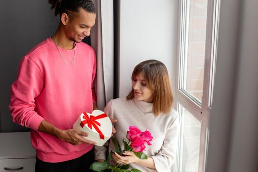 A stylish guy in a pink jumper gives his girlfriend, sitting by the window, a white heart-shaped box with a bow and a bouquet of roses