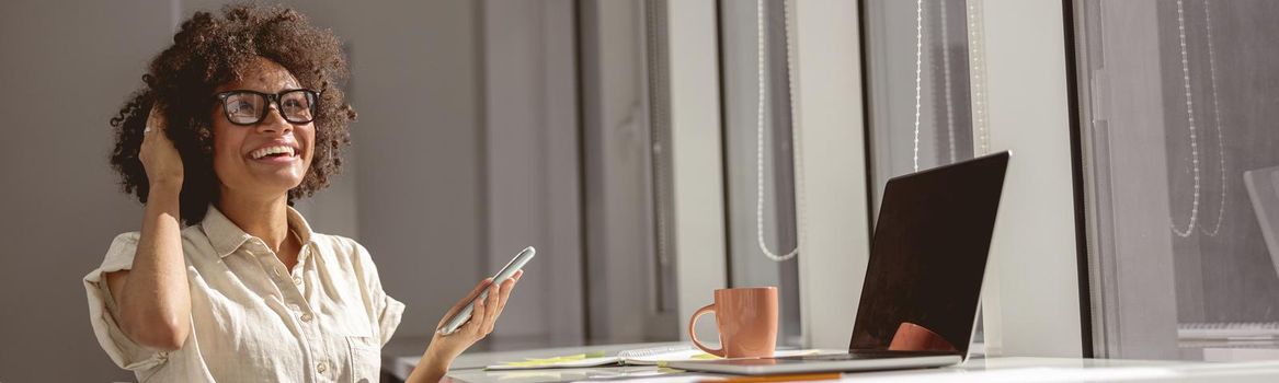 Happy young lady in headphones and holding mobile phone while sitting at her workplace