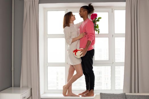 Multiracial modern, beautiful couple, a guy in a pink jumper and a girl in a white dress, stand on a large white window, hugging, holding a bouquet of roses and a gift box in the form of a heart