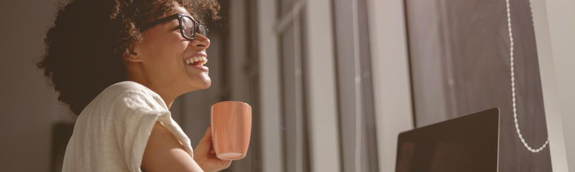 Happy young lady working with laptop and holding cup with drink in the office during sunny day