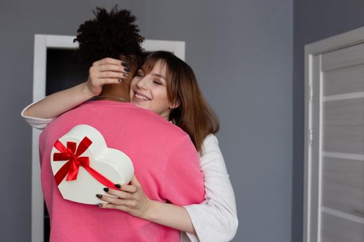 A happy beautiful girl, hugging her boyfriend in a pink jumper, holds a white box in the form of a heart with a bow in her hands