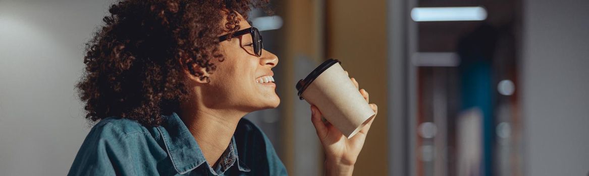 Smiling pretty woman in glasses drinking coffee while working in the modern office