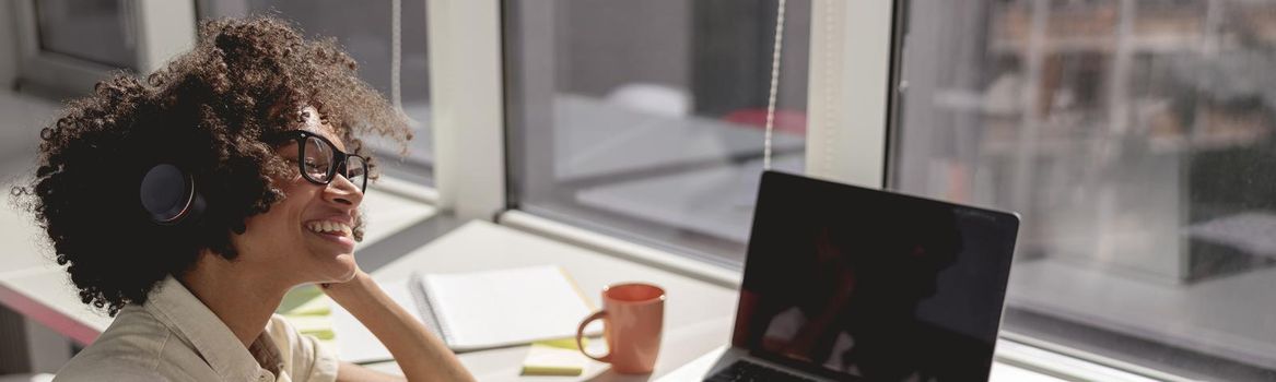 Pretty Afro American lady sitting at workplace with laptop while using headphones and smartphone in the office