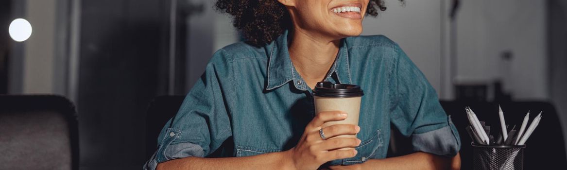 Smiling young multiethnic lady sitting at work desk and holding cup of coffee in the office