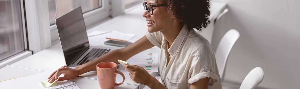 Side view of smiling woman working with laptop and notebook while sitting near the window