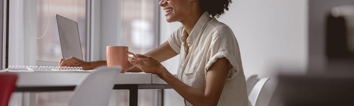 Side view of happy African American woman holding stickers and working with laptop near the window