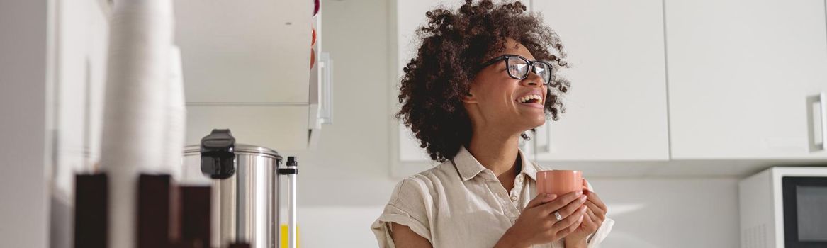 Pretty African American woman in glasses standing in modern office kitchen interior