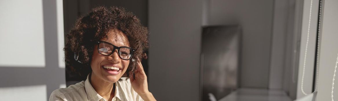 Pretty African American lady with headphones using laptop while holding documents at workplace