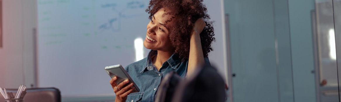 Happy Afro American businesswoman sitting at workplace and holding smartphone while looking away. Copy space