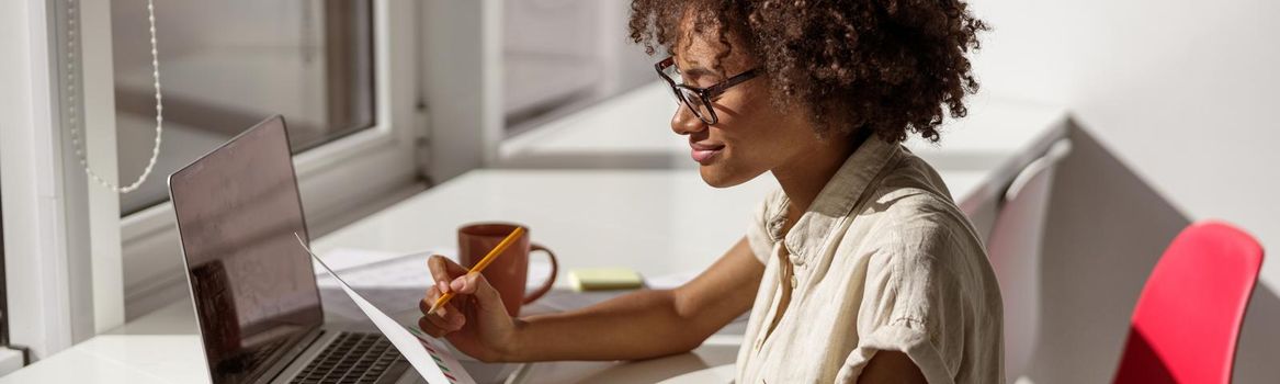 Happy pretty woman wearing glasses and sitting at workplace while holding document and reading