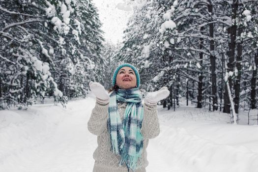 A girl in a white sweater, blue hat and scarf stands in the winter forest, cheerfully throws up the snow in white mittens