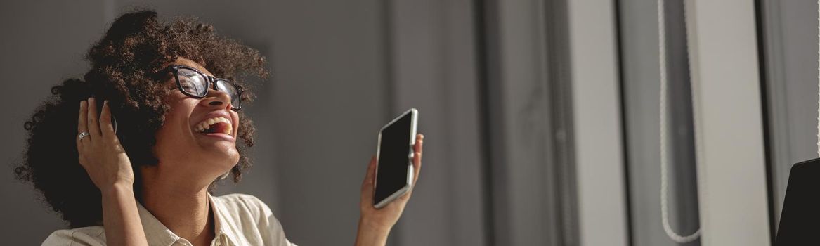 Happy African American female sitting in the coworking headphones and holding mobile phone