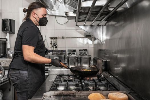 Chef in protective face mask preparing food in the kitchen of a restaurant or hotel during new normal. Coronavirus prevention concept. High quality photo