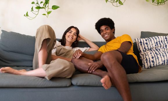 Smiling young interracial couple relaxing on the couch looking at camera. Lifestyle concept.