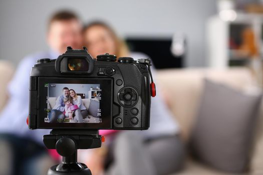 Close-up of camera stand on tripod with picture preview on screen, family posing on couch with kid. Self made photoshot at home. Family, memory concept