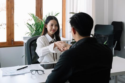 Two business people handshake at meeting table in office together with confident. Young asian businessman and businesswoman workers express agreement of investment deal.