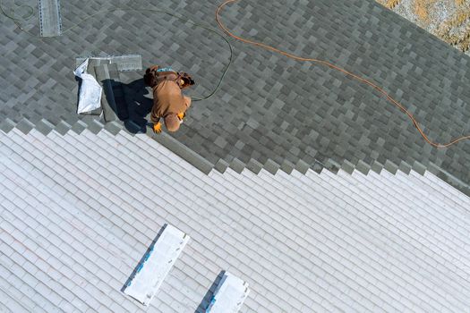 A worker installing shingles on the roof of a home with air hammer and nails
