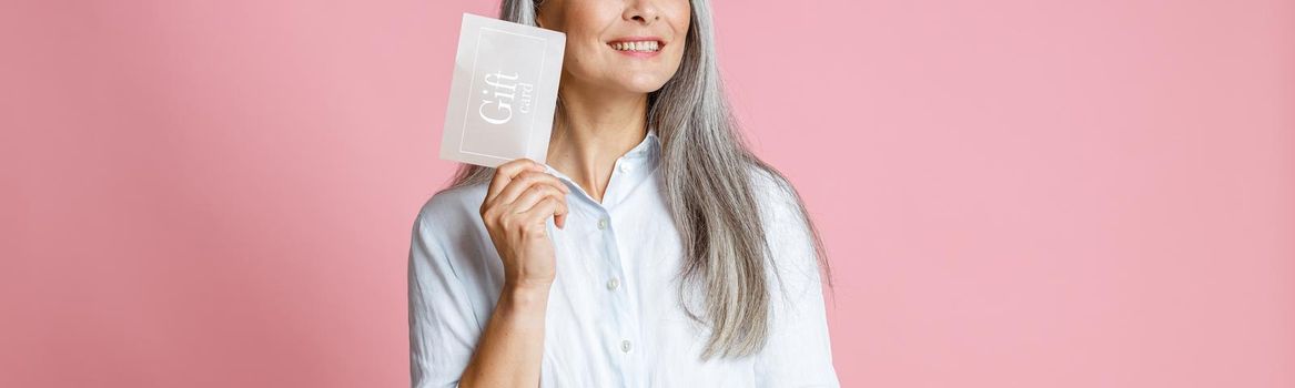 Pretty cheerful silver haired Asian woman holds gift card having ideas on pink background in studio. Shopping certificate