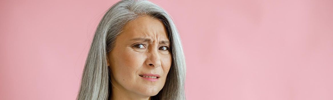 Disgusting grey haired Asian woman holds sonic facial cleansing brush on pink background in studio. Mature beauty lifestyle