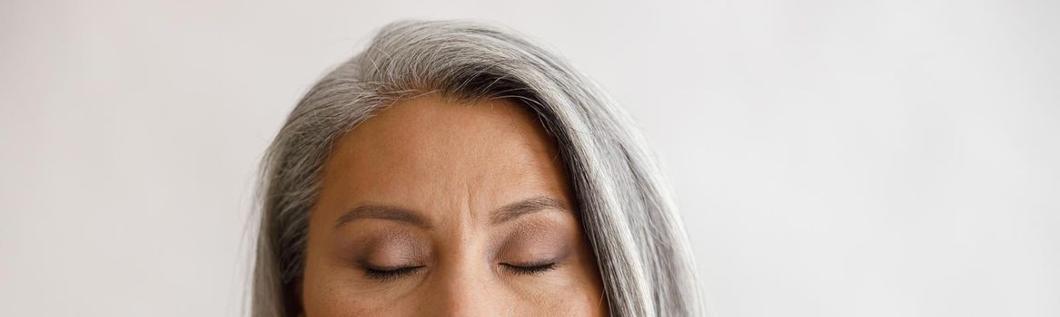 Pretty tranquil grey haired lady with closed eyes and elegant makeup poses on light background in studio. Mature beauty lifestyle