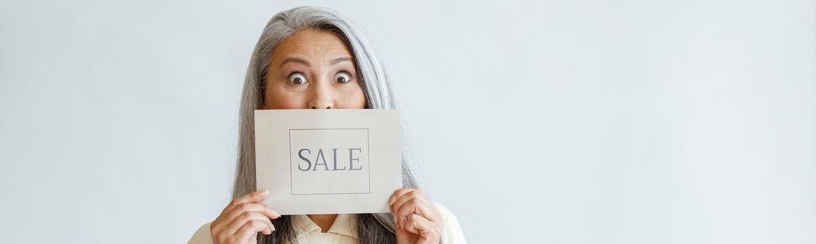 Shocked silver haired Asian lady holds card with word Sale on light grey background in studio, space for text