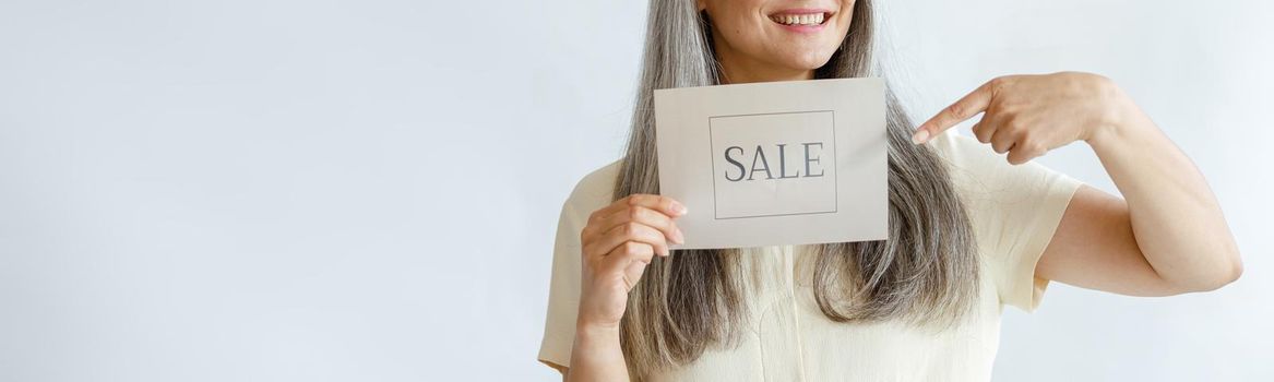 Joyful middle aged Asian woman with long grey hair points onto Sale sign posing on light background in studio, space for text