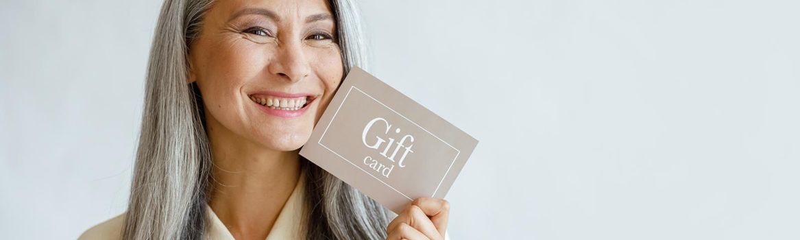 Pretty smiling mature Asian lady holds gift card posing on light grey background in studio, space for text. Shopping certificate