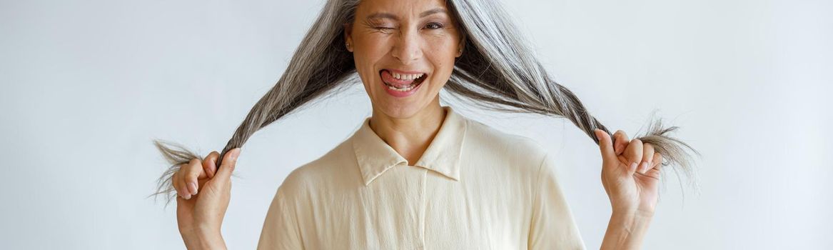 Funny middle aged Asian lady grimaces and holds long silver hair standing on light grey background in studio. Mature beauty lifestyle