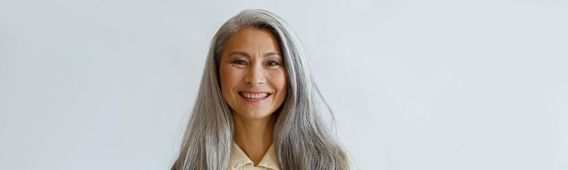 Portrait of smiling Asian woman with long hoary hair and crossed arms standing on light grey background in studio. Mature beauty lifestyle