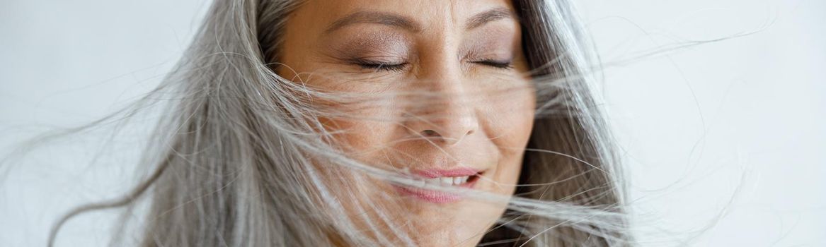 Happy Asian female model with flying hoary hair stands on light grey background in studio closeup. Mature beauty lifestyle