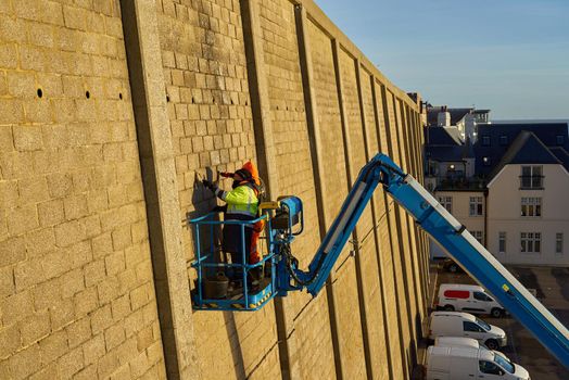 Two men on a blue boom arm make repairs to a tall cliff wall