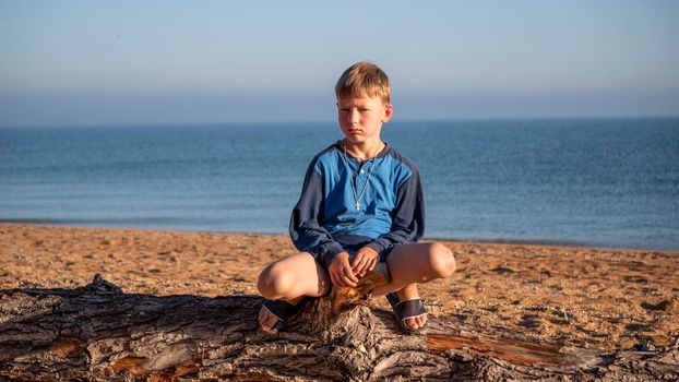 Portrait of a boy. A child is sitting on a log lying on the beach. A boy on the coast. Family vacation at the sea. Child on the sea.