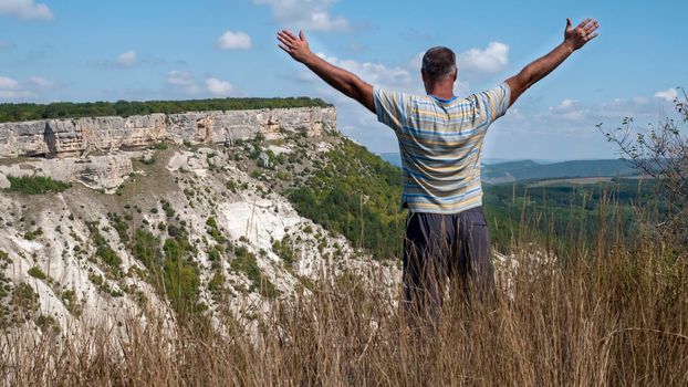 An adult man with his arms spread wide, seen from the back, stands on the edge of a precipice. Figure of a man on the background of a mountainous area. The man over the gorge.